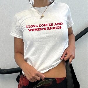 K&ouml;nnte beinhalten: Wei&szlig;es T-Shirt mit rotem Text "I LOVE COFFEE AND WOMEN'S RIGHTS". Das kurz&auml;rmelige Shirt scheint aus Baumwolle zu sein. Eine goldene Halskette und ein Ring sind sichtbar. Die Person h&auml;lt eine schwarze Tasche. Innenaufnahme.