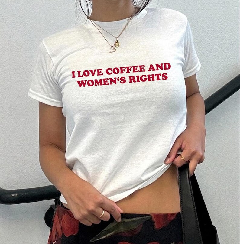 K&ouml;nnte beinhalten: Wei&szlig;es T-Shirt mit rotem Text "I LOVE COFFEE AND WOMEN'S RIGHTS". Das kurz&auml;rmelige Shirt scheint aus Baumwolle zu sein. Eine goldene Halskette und ein Ring sind sichtbar. Die Person h&auml;lt eine schwarze Tasche. Innenaufnahme.