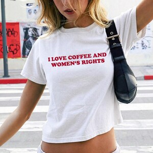 K&ouml;nnte beinhalten: Wei&szlig;es Crop-Top mit dem Text "I LOVE COFFEE AND WOMEN'S RIGHTS" in Rot. Das Shirt hat kurze &Auml;rmel und einen runden Ausschnitt. Auf der rechten Seite des Bildes ist eine schwarze Umh&auml;ngetasche zu sehen.