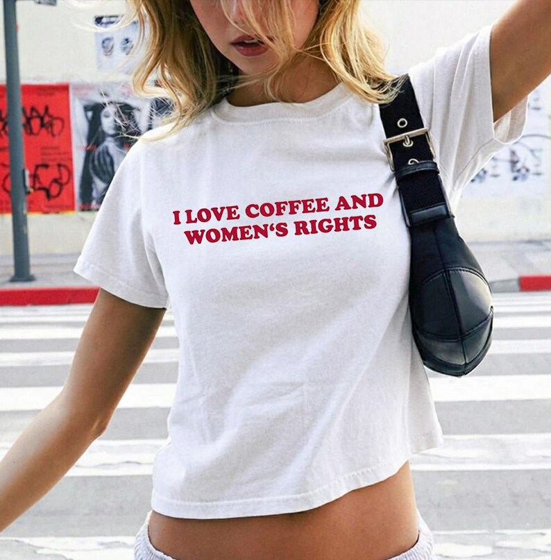 K&ouml;nnte beinhalten: Wei&szlig;es Crop-Top mit dem Text "I LOVE COFFEE AND WOMEN'S RIGHTS" in Rot. Das Shirt hat kurze &Auml;rmel und einen runden Ausschnitt. Auf der rechten Seite des Bildes ist eine schwarze Umh&auml;ngetasche zu sehen.