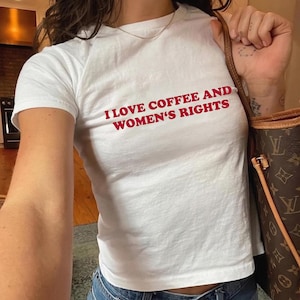 K&ouml;nnte beinhalten: Wei&szlig;es T-Shirt mit dem roten Text "I LOVE COFFEE AND WOMEN'S RIGHTS". Das Shirt hat kurze &Auml;rmel und scheint aus einem weichen Material zu sein. Die Person tr&auml;gt eine blaue Jeans und eine goldene Halskette.