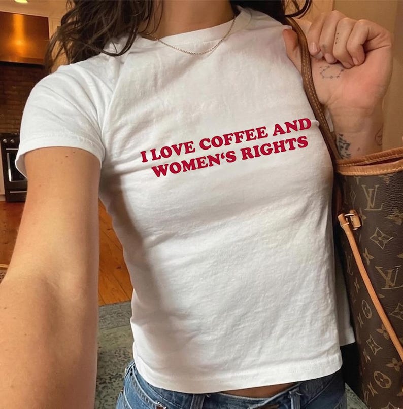 K&ouml;nnte beinhalten: Wei&szlig;es T-Shirt mit dem roten Text "I LOVE COFFEE AND WOMEN'S RIGHTS". Das Shirt hat kurze &Auml;rmel und scheint aus einem weichen Material zu sein. Die Person tr&auml;gt eine blaue Jeans und eine goldene Halskette.