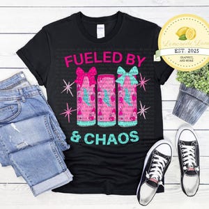 Może przedstawiać: Czarny T-shirt z napisem "FUELED BY" i trzema r&oacute;żowymi i niebieskimi puszkami z kokardkami. Napis "& CHAOS" znajduje się pod puszkami.
