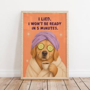 Op de afbeelding: Een ingelijste wandkunstprint met een gouden hond die een lavendelkleurige handdoek op het hoofd en een witte badjas draagt. De hond heeft komkommerschijfjes op de ogen en houdt een klein wit koffiekopje vast. De oranje achtergrond toont de tekst I LIED. I WON'T BE READY IN 5 MINUTES. met kleine witte sterren.