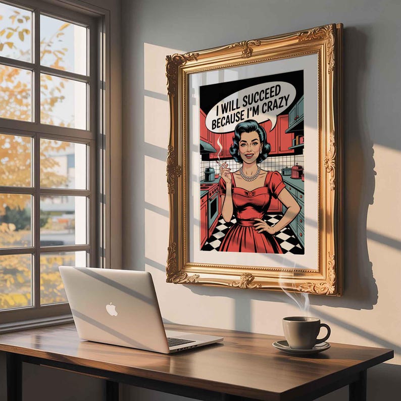 May include: A framed pop art print featuring a woman in a red dress and the text "I WILL SUCCEED BECAUSE I'M CRAZY" in a speech bubble. The print is in a gold ornate frame, hanging on a wall. A laptop and a cup of coffee sit on a wooden desk.