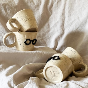 Può includere: Più tazze in ceramica macchiettata con dettagli neri. Una tazza ha occhiali neri, un'altra ha il testo "DON'T LET THE MUG GET YOU DOWN". Le tazze hanno una base color crema con macchie nere.
