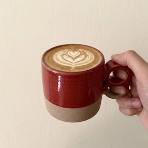 Peut inclure: Une tasse en céramique rouge avec une base beige, remplie d'un latte avec un motif en forme de cœur dans la mousse. La tasse est tenue par une main, mettant en valeur la conception unique de la poignée.