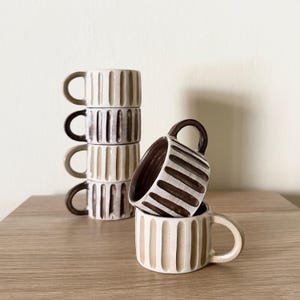 Pode incluir: Uma pilha de quatro canecas de cerâmica com textura canelada vertical e acabamento brilhante, apresentando um exterior creme e interior castanho escuro. Uma caneca a condizer está posicionada à frente, inclinada para mostrar o interior.
