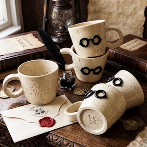 Può includere: Una collezione di tazze in ceramica maculata con dettagli di occhiali neri. Una tazza presenta il testo "DON'T LET THE MUGGLES GET YOU DOWN". Altri oggetti includono una penna, un calamaio, monete e una lettera di accettazione di Hogwarts.