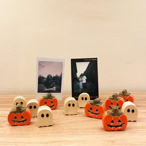 Puede incluir: Una colección de pequeños soportes de madera para tarjetas con temática de Halloween, que incluye calabazas naranjas con caras negras talladas y fantasmas de color blanco roto con ojos negros. Dos fotos Fujifilm Instax se muestran en los soportes.