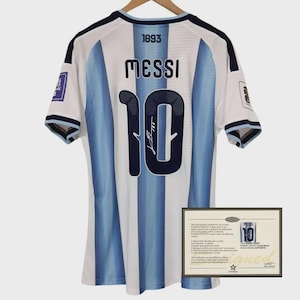 Puede incluir: Una camiseta a rayas blancas y azul claro con el nombre MESSI y el número 10 en azul oscuro en la parte posterior. El número 10 incluye una firma. El año 1893 está impreso sobre el nombre. Se incluye un certificado de autenticidad.