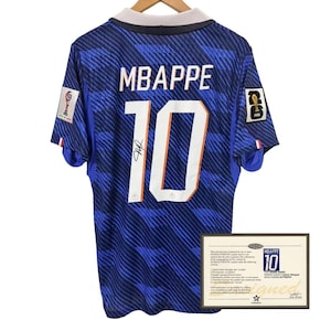 以下が含まれることがあります： 背中に「MBAPPE」の名前と「10」の番号が入った青いサッカーユニフォーム。ユニフォームは白い襟と番号の下にサインが入っています。真正性の証明書が下に表示されています。