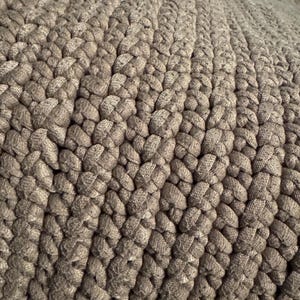 May include: Close-up of a woven textile in shades of gray and brown. The texture is created by a series of tightly knotted, thick cords, forming a pattern of raised, rounded shapes. The overall effect is a dense, textured surface.