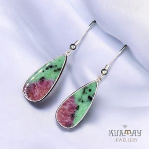 Op de afbeelding: Een paar druppelvormige oorbellen met een zilverkleurige metalen zetting. De oorbellen hebben een levendige combinatie van groene en rood-paarse edelstenen. De edelstenen hebben een gespikkelde uitstraling. De oorbellen worden op een witte ondergrond gepresenteerd.
