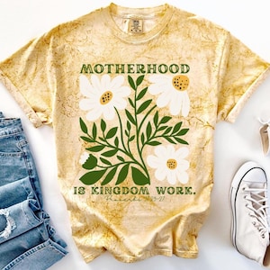 Puede incluir: Camiseta amarilla teñida con la frase "MOTHERHOOD IS KINGDOM WORK" y un diseño floral. El diseño presenta margaritas blancas con hojas y tallos verdes. La camiseta se combina con vaqueros azules y zapatillas blancas.