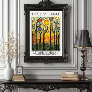 May include: A framed art print with a stained-glass design, featuring the text "GUSTAV KLIMT" and "MUSEE D'ORSAY". The artwork is in a black ornate frame, displayed on a black table with a lamp and vase. The print has a colorful design.