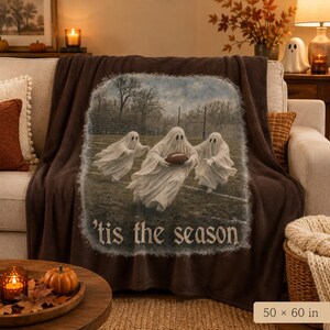May include: A dark brown throw blanket featuring a central graphic of three white ghosts playing American football on a field. The text 'tis the season' is printed below the ghosts. The blanket measures 50 inches by 60 inches in total size.