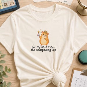 May include: A cream-colored t-shirt featuring a graphic of a hamster wearing a crown and holding a star-tipped wand. Below the hamster, the text reads: for my next trick... the disappearing lisp. The shirt has a crew neckline and short sleeves.