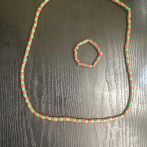May include: A necklace and bracelet set featuring alternating red and green beads. The necklace is a long strand, and the bracelet is a smaller, circular design. The jewelry is displayed on a dark wooden surface.