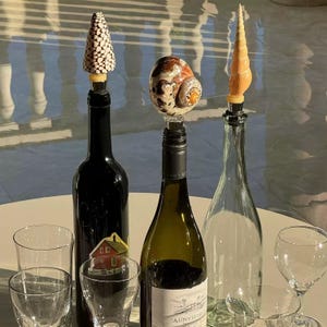 May include: Three wine bottles with decorative stoppers. One stopper is a cone shape with white and brown spots, another is a seashell, and the third is a spiral seashell. Clear wine glasses are in the foreground.