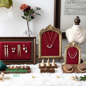 May include: Jewellery display featuring necklaces, earrings, and rings. The items are presented on wooden and gold display stands with a red velvet background. The scene is set on a white, fluffy surface, with a vintage aesthetic.