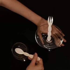 以下が含まれることがあります： 白いフォークと白いスプーンが付いた2つの透明なグラス。フォークとスプーンは装飾的で波状のデザインです。背景は黒です。