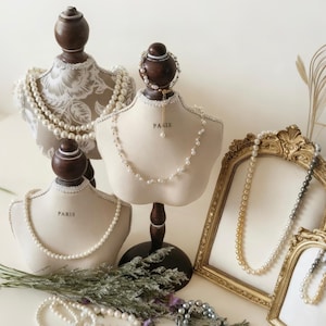 May include: An arrangement of jewelry display busts with pearl necklaces and bracelets. The busts are cream-colored with wooden stands and tops. One bust has the word "PARIS" printed on it. Gold-framed mirrors display additional pearl necklaces.
