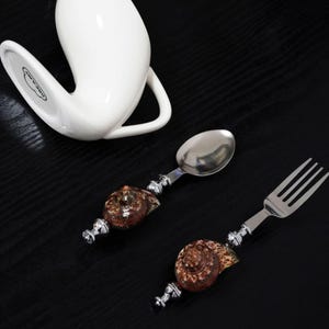 May include: A stainless steel spoon and fork set with decorative handles. The handles feature a brown and tan seashell design with silver-colored accents. A white ceramic mug is in the background.