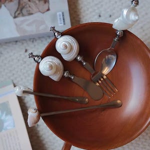 Puede incluir: Un conjunto de cubiertos únicos con mangos de concha, que incluye cucharas, tenedores y cuchillos, dispuestos en un cuenco de madera. Los mangos están adornados con detalles metálicos plateados. El cuenco es de color marrón cálido.