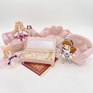 Puede incluir: Una caja de joyería rosa con espejo, pulseras y anillos dorados descansa sobre una pequeña alfombra roja. Sofás rosas en miniatura y muñecas están dispuestas alrededor de la caja de joyería, creando una escena caprichosa. También hay una pequeña figura de perro.