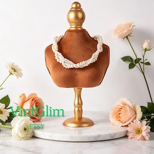 May include: A gold-colored jewelry display stand with a brown velvet bust, showcasing a white pearl necklace. The stand is adorned with a gold finial and base, set against a backdrop of flowers.