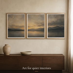 Könnte beinhalten: Drei gerahmte Landschaftsdrucke mit einem Sonnenuntergang über einem ruhigen See und fernen Bergen in hellen Holzrahmen. Unter der Kunst steht der Text: Art for quiet interiors, PIXELVISONEU, DIGITAL ART FOR QUIET INTERIORS.