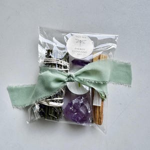 May include: An energy cleansing set in a clear cellophane bag, tied with a sage green ribbon. The set includes a bundle of sage, amethyst crystals, palo santo, and a small candle. The label reads "NOEMA ENERGY CLEANSING SET."