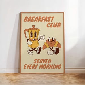 Puede incluir: Un póster enmarcado con un diseño de dibujos animados retro. La imagen presenta una cafetera y un cruasán, ambos con caras y extremidades animadas. El texto dice "BREAKFAST CLUB SERVED EVERY MORNING" en letras marrones sobre un fondo crema.
