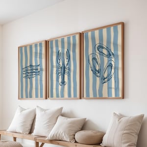 Può includere: Tre stampe artistiche incorniciate con illustrazioni di frutti di mare. Ogni stampa ha uno sfondo a righe blu e crema. Le stampe raffigurano pesci, un'aragosta e ostriche in uno stile di disegno a linee blu. Le cornici sono in legno.