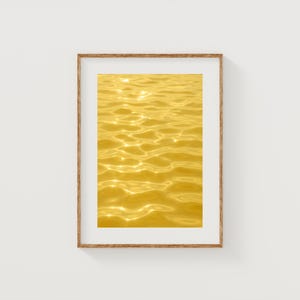 Puede incluir: Impresión enmarcada que muestra un primer plano de ondulaciones doradas en el agua. La superficie del agua refleja la luz solar, creando un efecto brillante. La obra de arte se exhibe en un marco de madera.