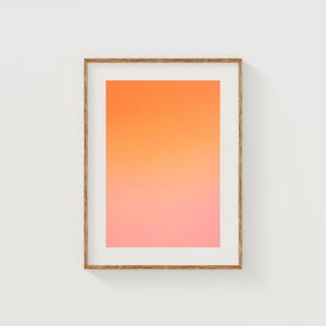 Puede incluir: Una impresión artística enmarcada que presenta un degradado de colores, pasando del naranja en la parte superior al rosa en la parte inferior. La obra de arte está encerrada en un marco de madera marrón claro, creando una estética moderna. La impresión está montada en una pared blanca.