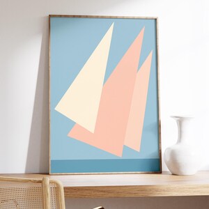 May include: Framed abstract art print featuring geometric shapes in pastel colors. The artwork displays three triangular shapes in cream and peach against a light blue background. A horizontal blue line is at the bottom. The frame is a light brown color.