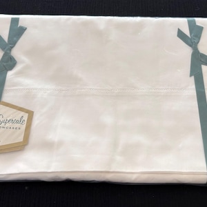 May include: A rectangular package of white Wamsutta Supercale sheets and pillowcases measuring 72 x 108 inches. The folded bedding is wrapped in clear plastic and secured with two teal ribbons tied in bows, featuring a tan hexagonal product tag.