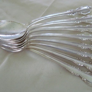 May include: A collection of silver spoons arranged in a fan shape. The spoons have a reflective, polished surface and ornate detailing on the handles. The spoons are likely for dining and are set against a neutral background.