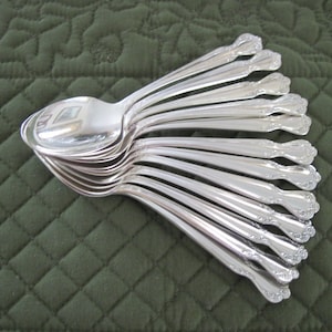 May include: A collection of shiny, silver spoons arranged in a fan shape. The spoons have a decorative floral pattern on the handles and are resting on a dark green, quilted surface. The spoons are made of a reflective metal.