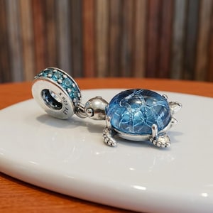 Könnte beinhalten: Ein Schildkröten-Charm aus Sterlingsilber mit einem gewölbten, blauem Glaspanzer mit Rissmuster. Die Öse ist mit kleinen blauen Kristallen besetzt und mit dem Text S 925 ALE graviert. Die Schildkröte hat einen Kopf und Flossen aus poliertem Silber.
