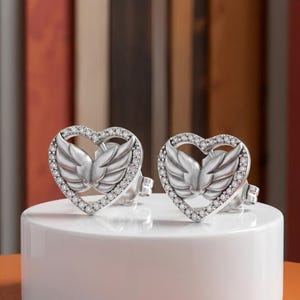 May include: A pair of silver-coloured heart-shaped stud earrings. Each earring features a pair of detailed wings within the heart outline, surrounded by small, sparkling stones. The earrings are displayed on a white surface.