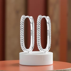 May include: A pair of silver oval hoop earrings. The earrings feature a textured, braided pattern along the outer edge. Each earring post is stamped with "S925". The earrings are presented on a white plinth against a blurred backdrop.