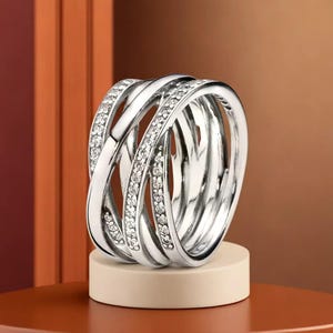 May include: A silver ring with an intricate, interwoven design. The ring has multiple bands that cross over each other, with one band set with small, sparkling stones. The ring is displayed on a small, off-white pedestal.