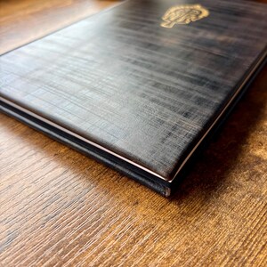 Puede incluir: Un cuaderno rectangular con una cubierta texturizada de color gris carb&oacute;n oscuro que presenta un emblema dorado en relieve en la parte superior. El cuaderno est&aacute; cerrado, mostrando un borde blanco fino de las p&aacute;ginas entre las cubiertas.