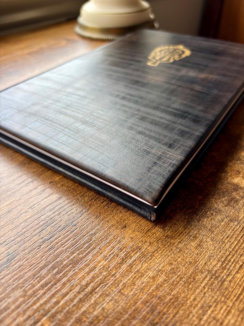 Puede incluir: Un cuaderno rectangular con una cubierta texturizada de color gris carb&oacute;n oscuro que presenta un emblema dorado en relieve en la parte superior. El cuaderno est&aacute; cerrado, mostrando un borde blanco fino de las p&aacute;ginas entre las cubiertas.