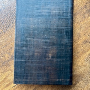 Puede incluir: Un libro rectangular con una cubierta de color negro oscuro que presenta una textura entrecruzada visible. La superficie muestra sutiles variaciones de tono, revelando toques de marr&oacute;n bajo el acabado negro, creando un aspecto desgastado.