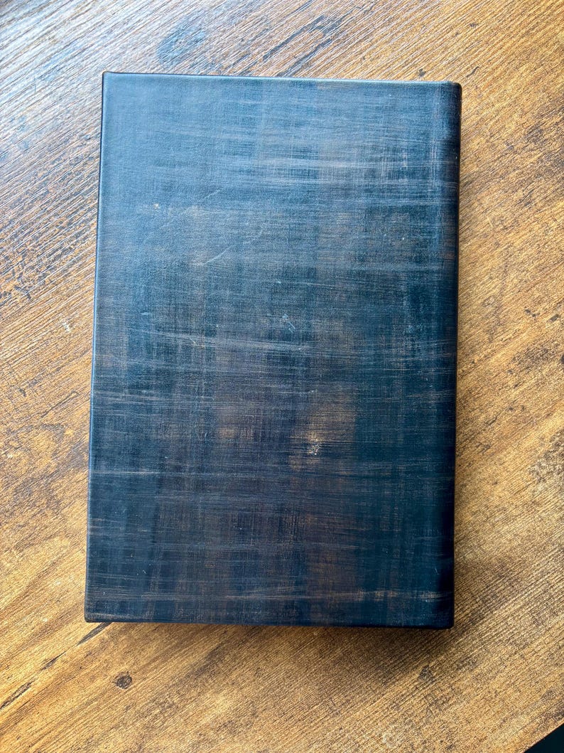Puede incluir: Un libro rectangular con una cubierta de color negro oscuro que presenta una textura entrecruzada visible. La superficie muestra sutiles variaciones de tono, revelando toques de marr&oacute;n bajo el acabado negro, creando un aspecto desgastado.