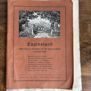 May include: A vintage book cover titled "Euginsland" with a sepia-toned illustration of a landscape scene. The cover features German text and a list of locations. The book is bound with a light brown cover and has a worn appearance.